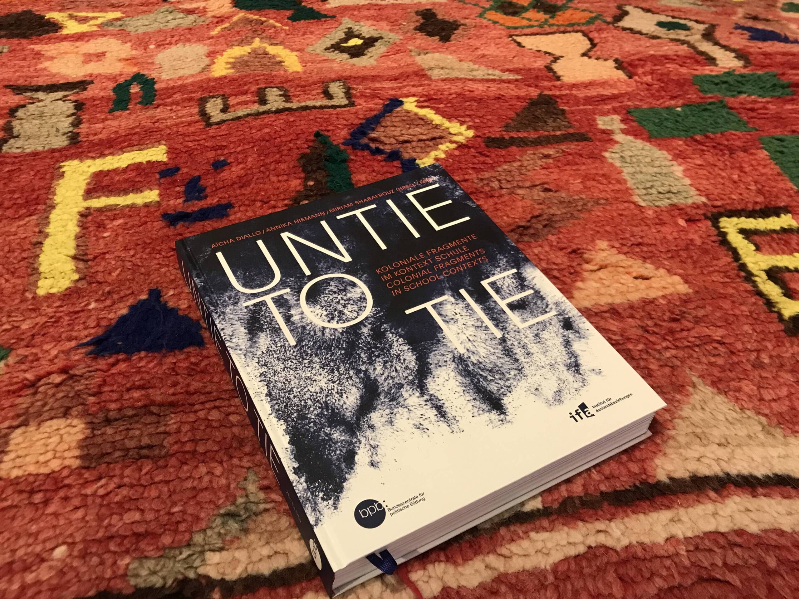 Das Buch Untie to Tie. Koloniale Fragmente im Kontext Schule liegt auf einem marokkanischen Teppich